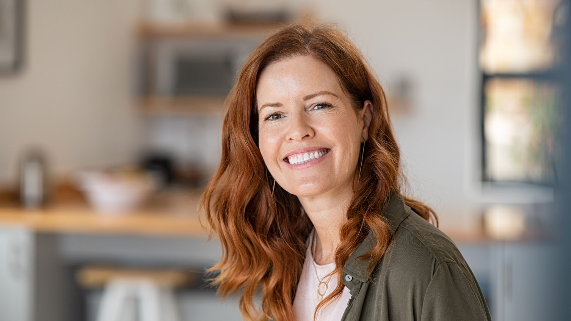 Smiling middle-aged woman with auburn hair wearing a green shirt and gold jewellery, standing in a kitchen